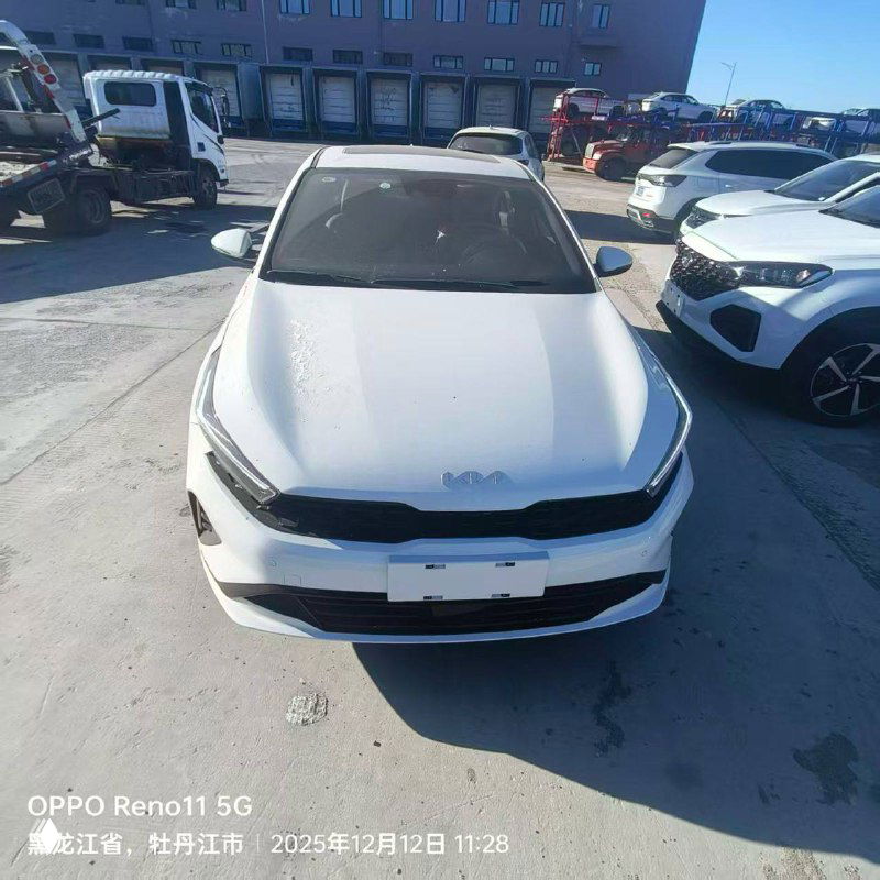 Белый KIA K3 спереди на торговой площадке в Китае: машина на ровной поверхности рядом с другими автомобилями и служебной техникой.