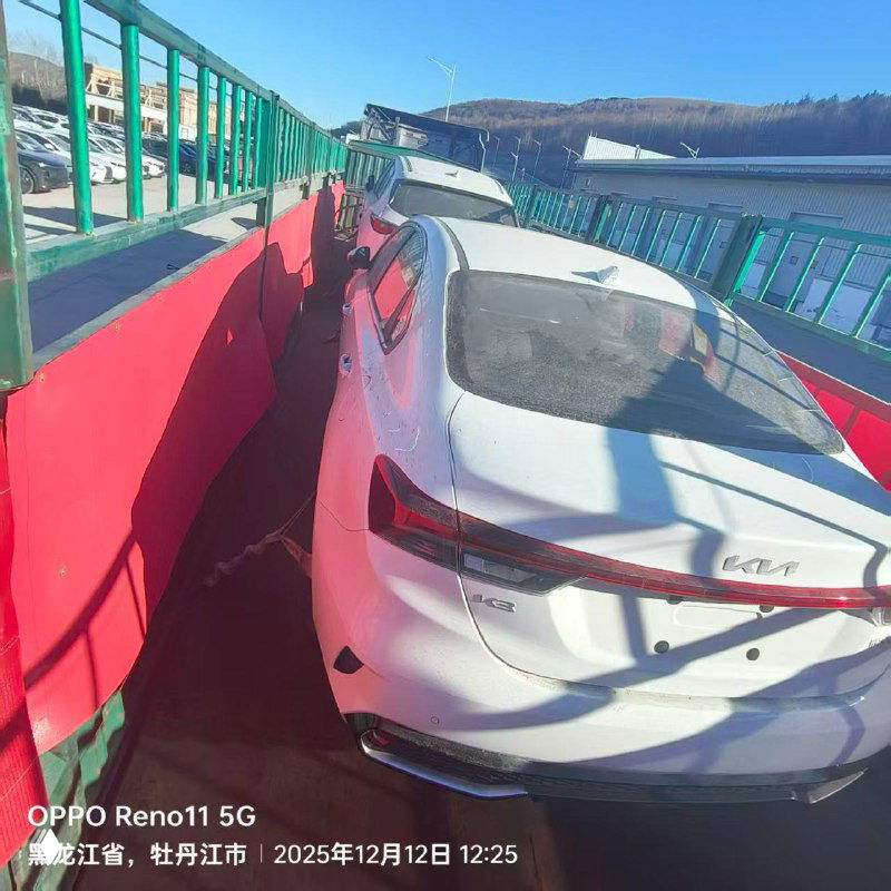 Вид сзади белого автомобиля на автовозе/площадке погрузки: багажник и фон с ограждением, фото сделано в дневное время.