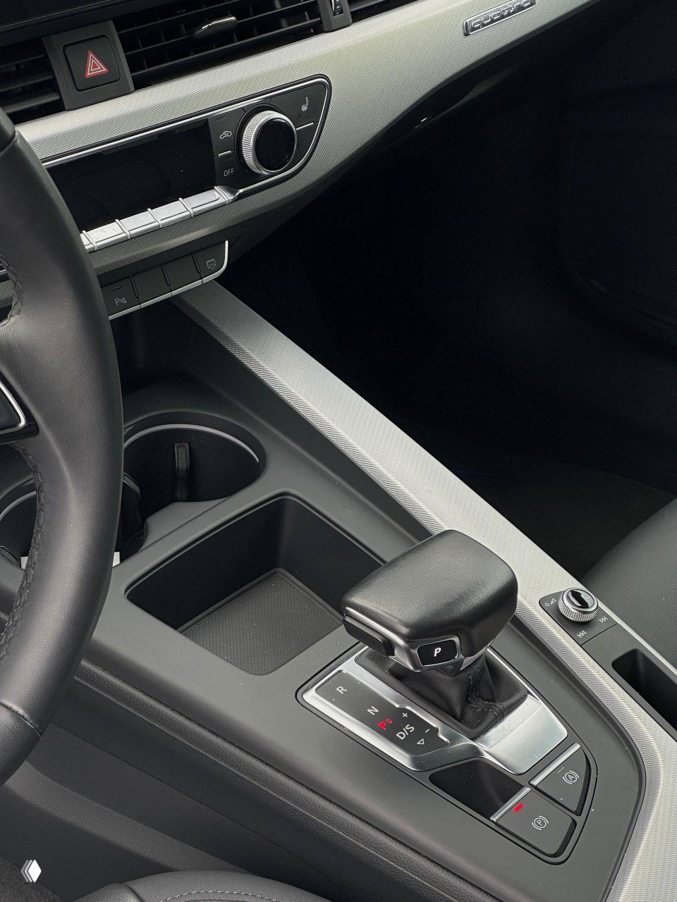 Крупный план центральной консоли и селектора коробки передач Audi, видны подстаканники, кнопки и отделка салона чёрного цвета