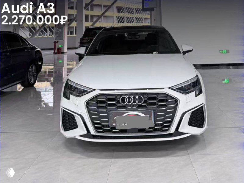 Фронтальный вид белого Audi A3 в выставочном зале: чётко видна передняя оптика, эмблема и название модели с указанием цены на изображении.