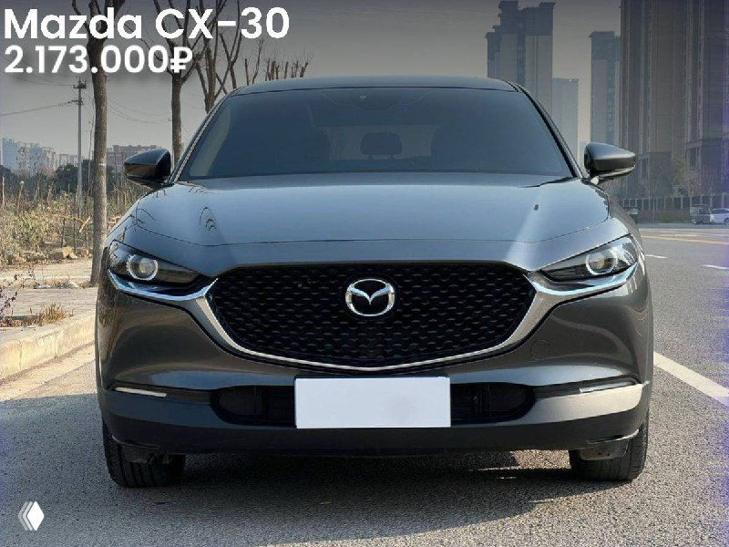 Фронтальное фото тёмной Mazda CX-30 2021 года на дороге в городской среде; видна маска радиатора, фары и указание цены в верхней части кадра.