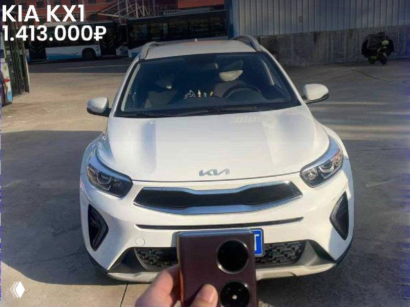 Фронтальный снимок белой KIA KX1 2021 года на площадке, видна передняя часть автомобиля и держимое в руке устройство внизу кадра.