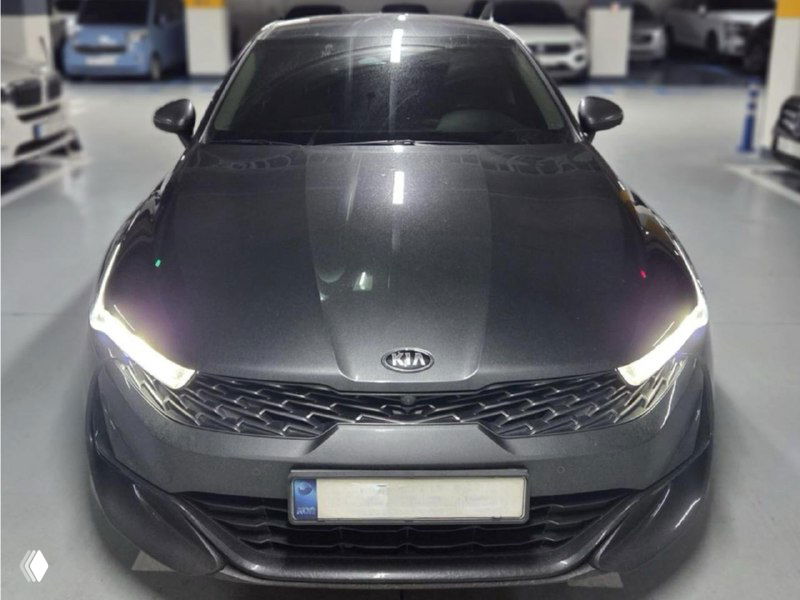 Фронтальный вид серой KIA K5 в гаражном помещении, вид на капот и решётку радиатора, фары включены, номерной знак размыто.