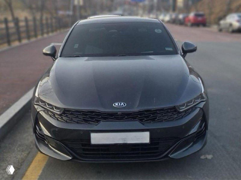 KIA K5 серого цвета, припаркованная у бордюра на улице: вид спереди, отражения на капоте и чистая облицовка бампера хорошо видны.