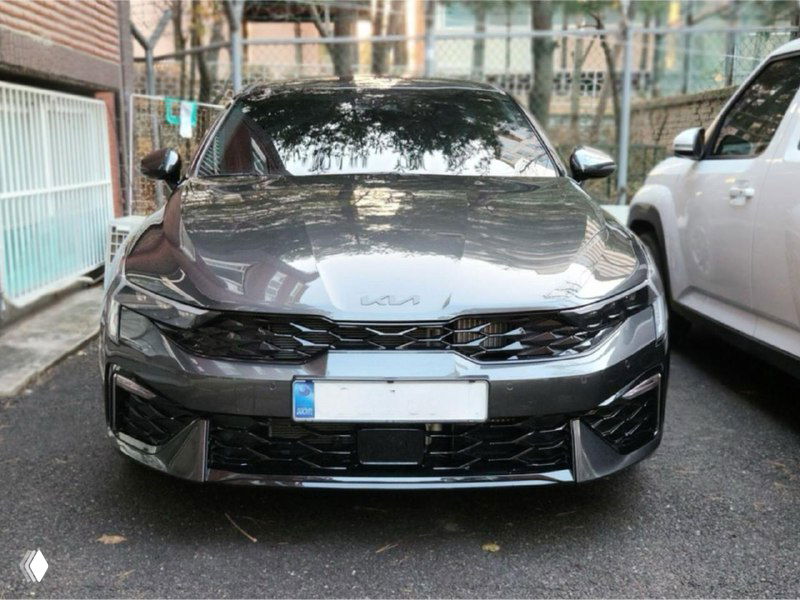 KIA K5 серого оттенка во дворе среди других машин, передняя часть машины под углом, видна решётка радиатора и форма капота.