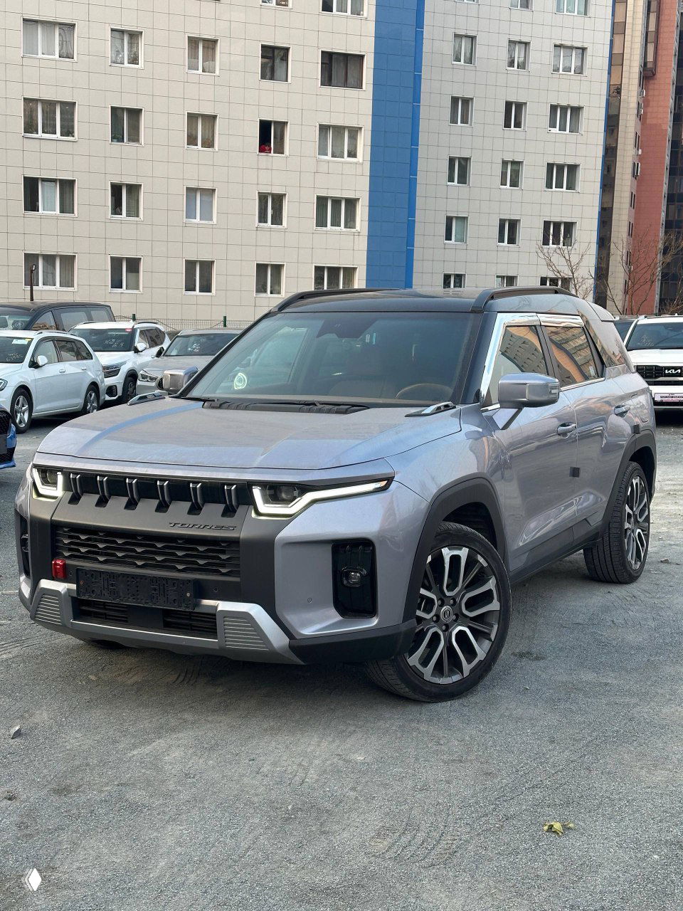 Внешний вид SsangYong Torres на стоянке, серый кузов, передний ракурс с агрессивной решёткой и дизайнерскими светодиодными фарами, автомобиль на фоне домов.