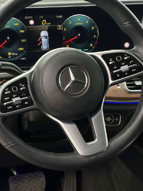 Крупный план рулевого колеса Mercedes GLE 450 с фирменной эмблемой, вид на цифровую приборную панель и элементы управления на спицах руля.