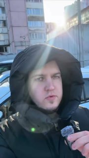 Человек в тёплой куртке на стоянке автомобилей зимой, держит микрофон; на заднем плане здания и солнечный блик — общий контекст обзора стоянки