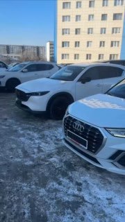 Ряд стоящих автомобилей на заснеженной парковке, на переднем плане белые кроссоверы (Mazda/Audi) и городской жилой корпус на заднем плане