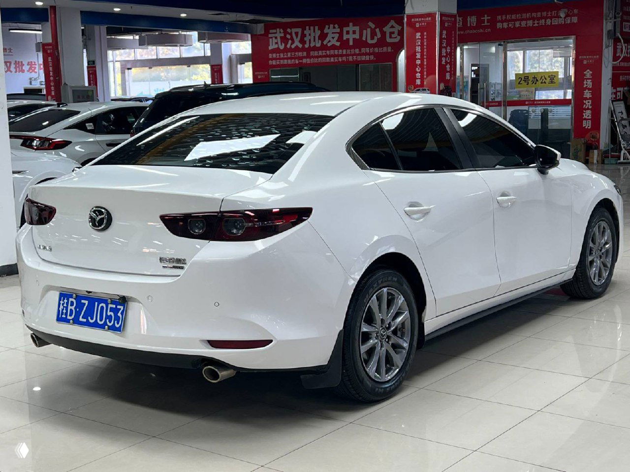 Белая Mazda 3 Axela, вид сзади в автосалоне: задние фонари, бампер, видна выхлопная труба и синяя китайская номерная табличка в помещении магазина.