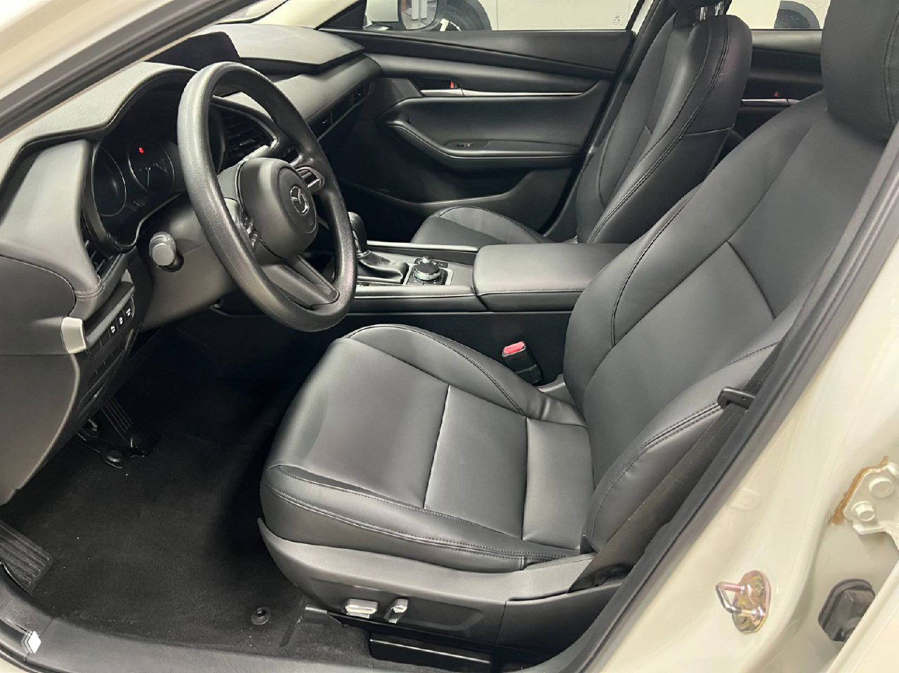Водительское место Mazda 3 Axela: чёрное кожаное сиденье, руль, подлокотник и панель управления — видно чистый и аккуратный салон с улицей шоурума на заднем плане.