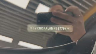 Кадр с фокусом на лобовом стекле Mazda 3 Axela: видна VIN-пластинка за стеклом и отражение рук фотографа, кадр из видеозаписи в автосалоне.