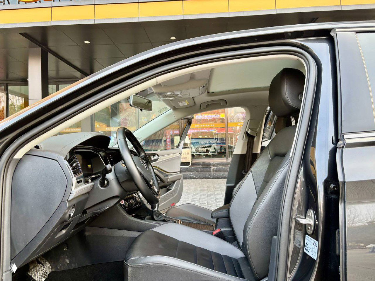 Вид на переднее пассажирское сиденье Volkswagen Sagitar: кожаная отделка, панорамная крыша сверху и открытая дверь на фоне автосалона.