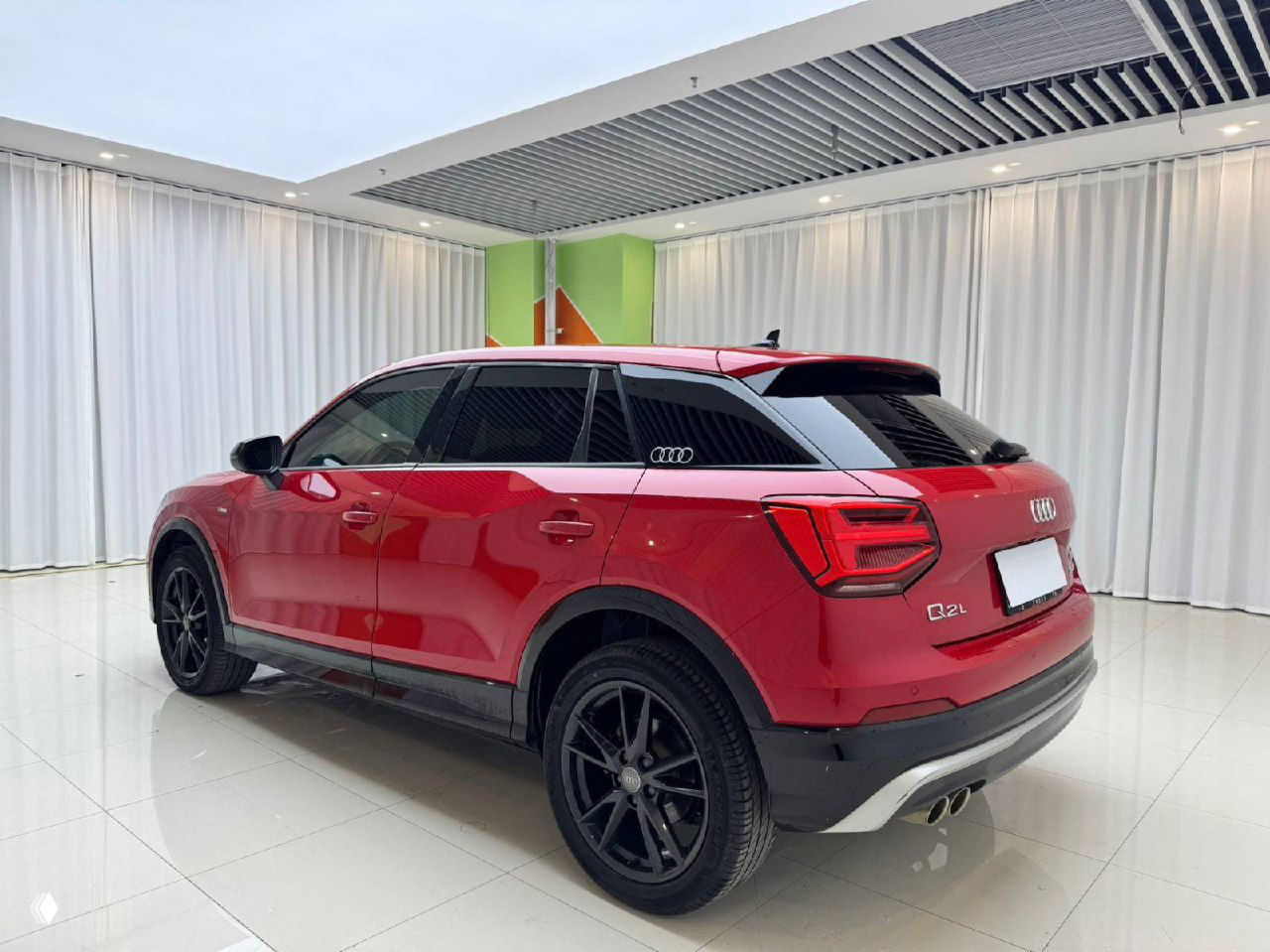 Красный Audi Q2L снятый с заднего диагонального ракурса в шоуруме, видна задняя оптика, черные диски и общая сохранность кузова без окрасов.