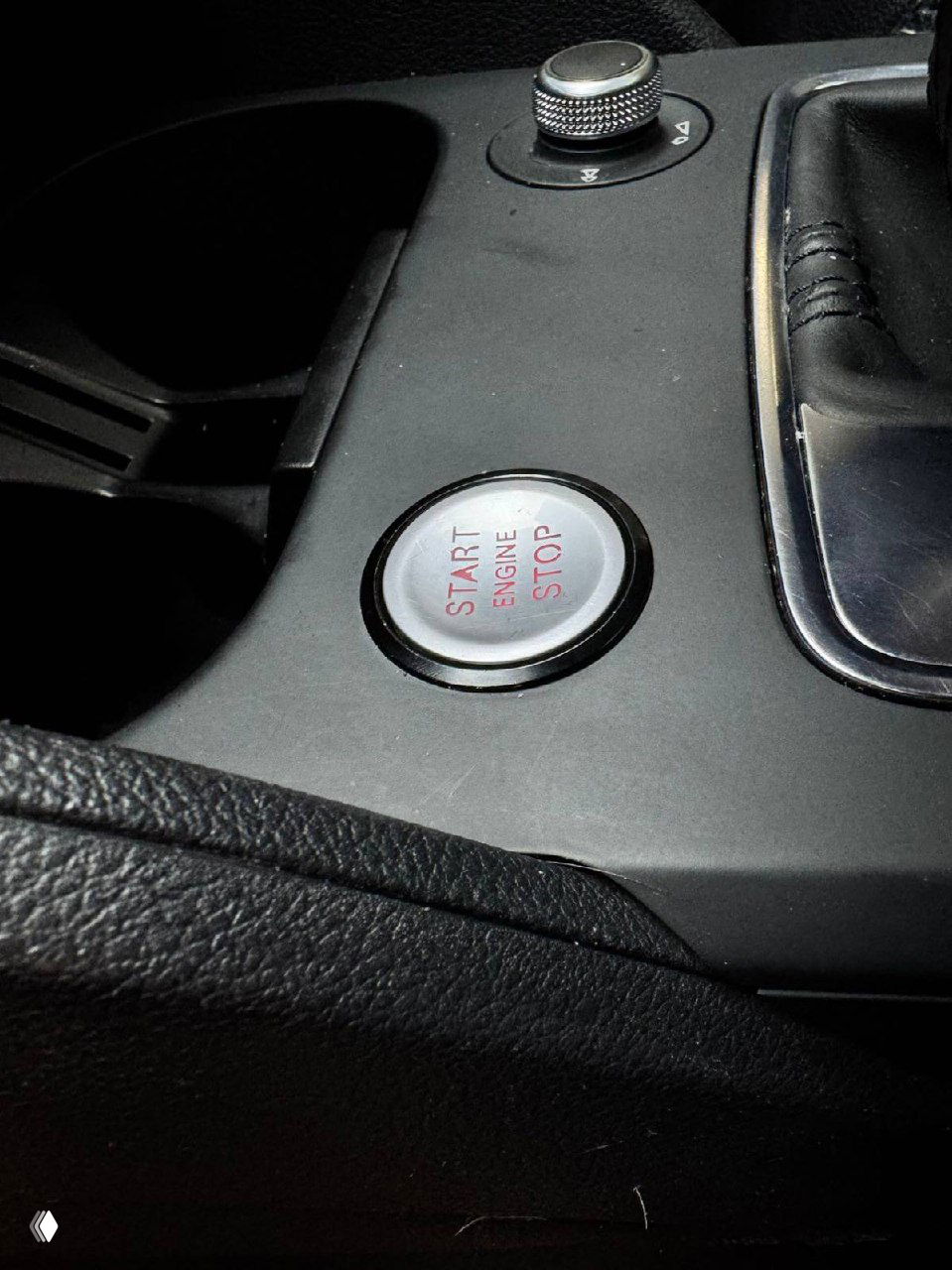 Крупный план кнопки Start/Stop внутри салона Audi Q2L, круглая металлическая кнопка с надписью START ENGINE STOP на чёрной панели приборов.