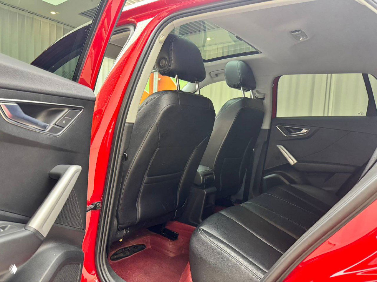 Вид на задние сиденья Audi Q2L с открытой передней дверью, чёрная обивка и чистый салон, видно панорамную крышу и подголовники пассажиров.