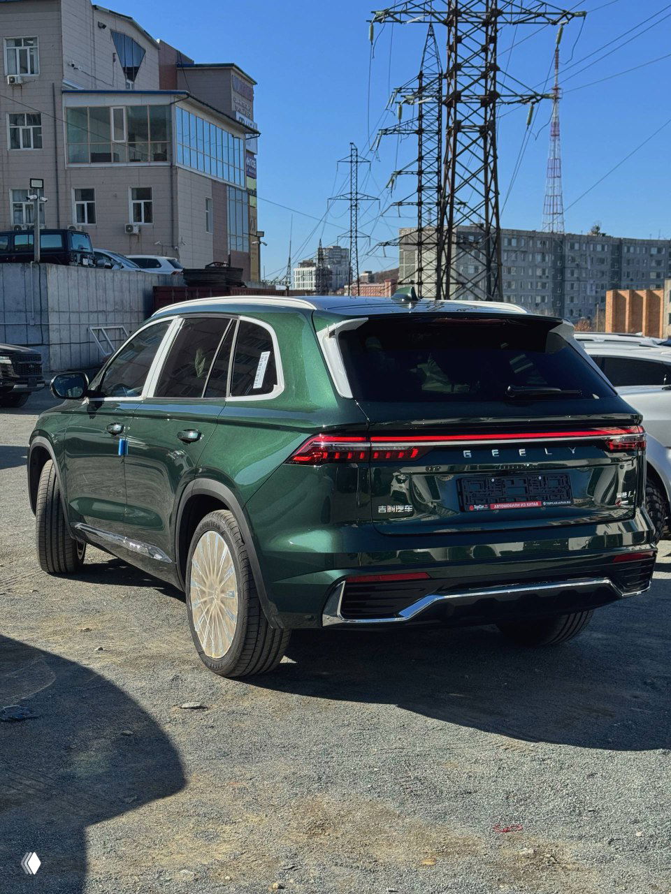 Geely Monjaro зелёного цвета сзади на стоянке, видна светодиодная световая полоса, эмблема GEELY и номерной модуль, фото сделано на городской площадке.