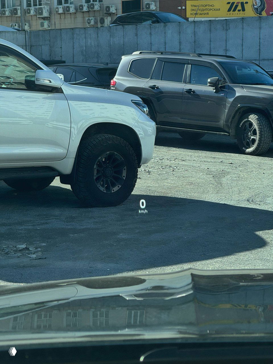 Вид с места водителя на стоянке с показанием 0 км на лобовом дисплее и автомобилями на фоне, демонстрируется нулевая пробеговая индикация и окружение стоянки.