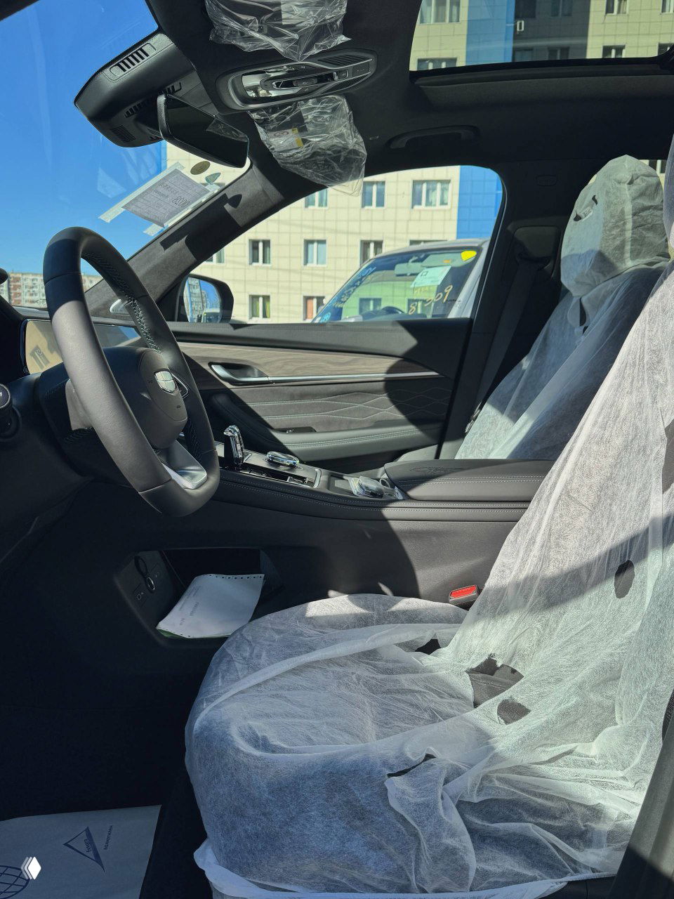 Передняя часть салона Geely Monjaro: руль, пассажирское кресло в защитной плёнке и элементы декоративной отделки торпедо на дневном освещении стоянки.