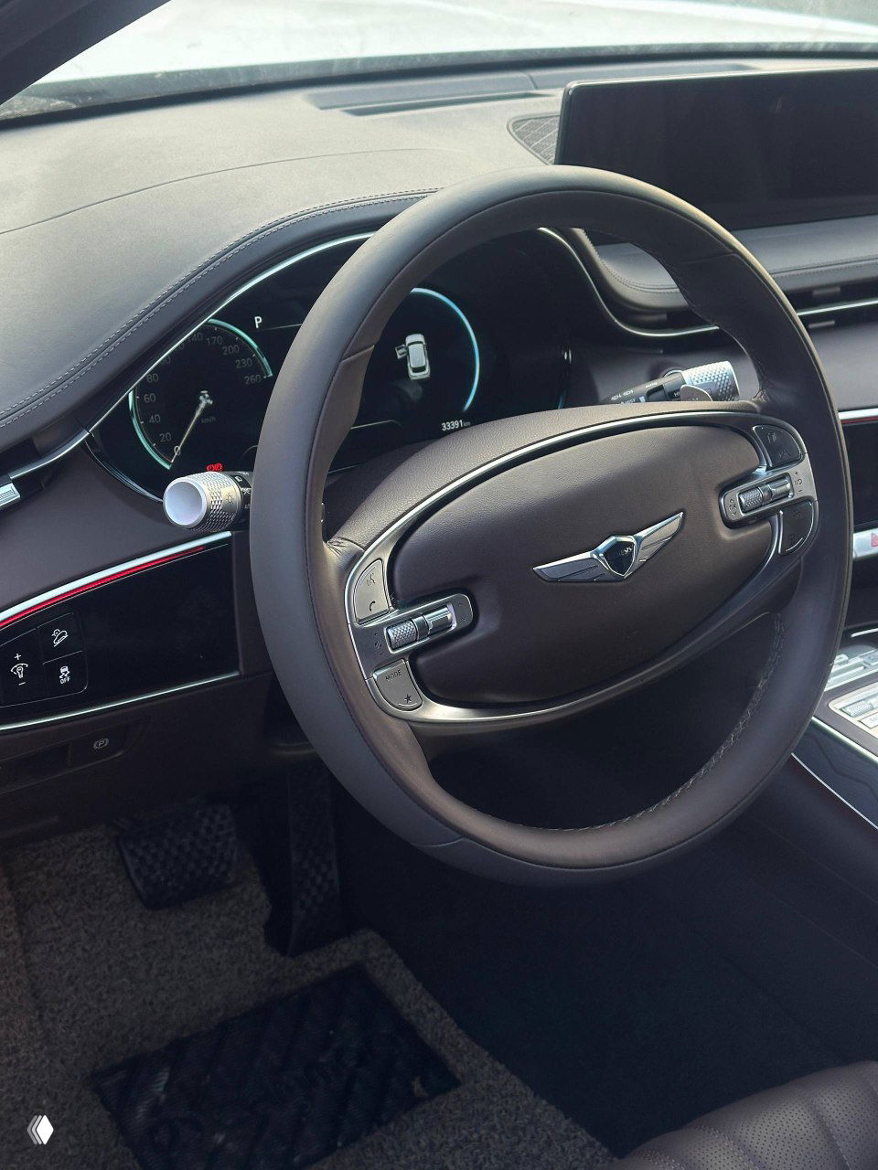 Крупный план рулевого колеса и приборной панели Genesis GV70: коричневый руль, цифровая приборка и элементы управления, видим пробег и отделку салона высокого класса.