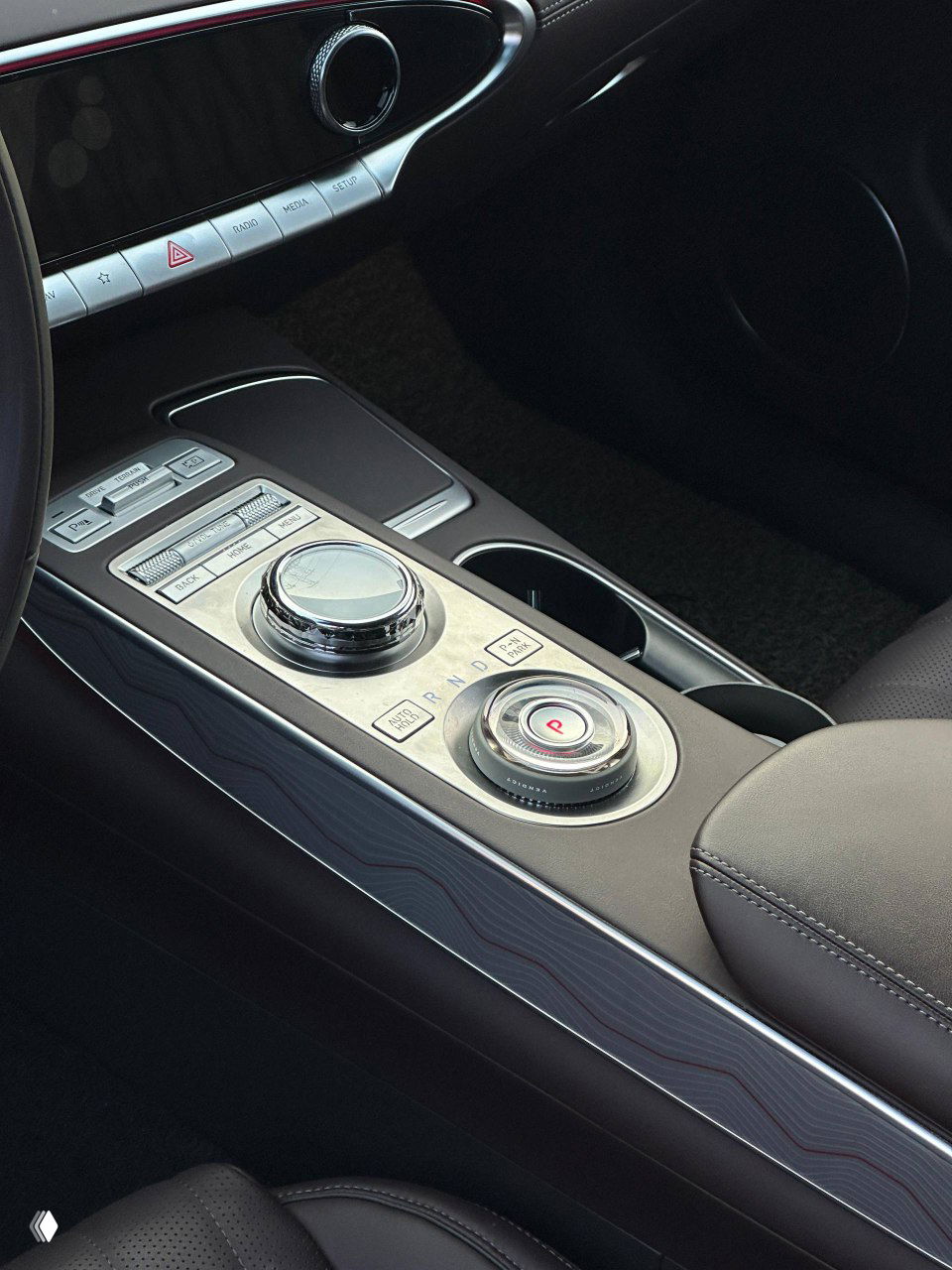 Деталь центральной консоли Genesis GV70: шайба селектора передач, кнопки управления климатом и медиасистемой, хромированные вставки и текстура отделки возле подстаканников.