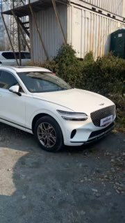 Вертикальная фотография белого Genesis GV70 в профиль на стоянке; видна боковая линия кузова, зеркала и задняя часть автомобиля на фоне контейнерной зоны и растительности.