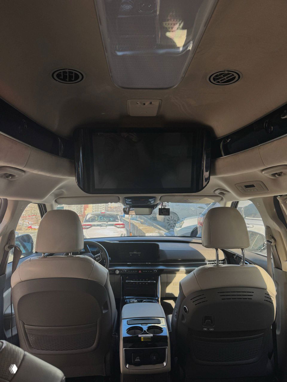 Салон KIA Carnival с опущенным потолочным монитором для задних пассажиров, вид на спинки передних кресел и центральную консоль сзади.