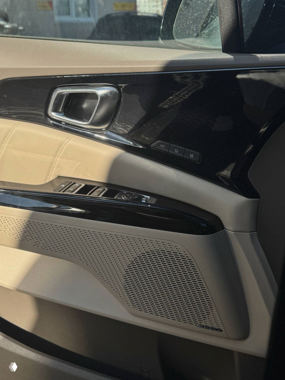 Крупный план дверной панель KIA Carnival — рукоять, кнопки управления стеклами и декоративная накладка, видна текстура материалов и динамик.