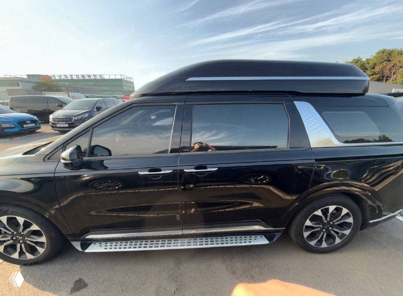 Боковой ракурс чёрного KIA Carnival на стоянке, видны боковые пороги, сдвижные двери и багажный бокс на крыше в контексте подготовки к транспортировке