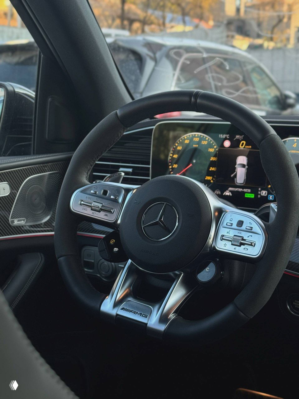 Руль Mercedes GLE53 AMG с логотипом AMG, мультифункциональные кнопки и цифровая приборная панель на фоне переднего стекла и паркующихся машин.