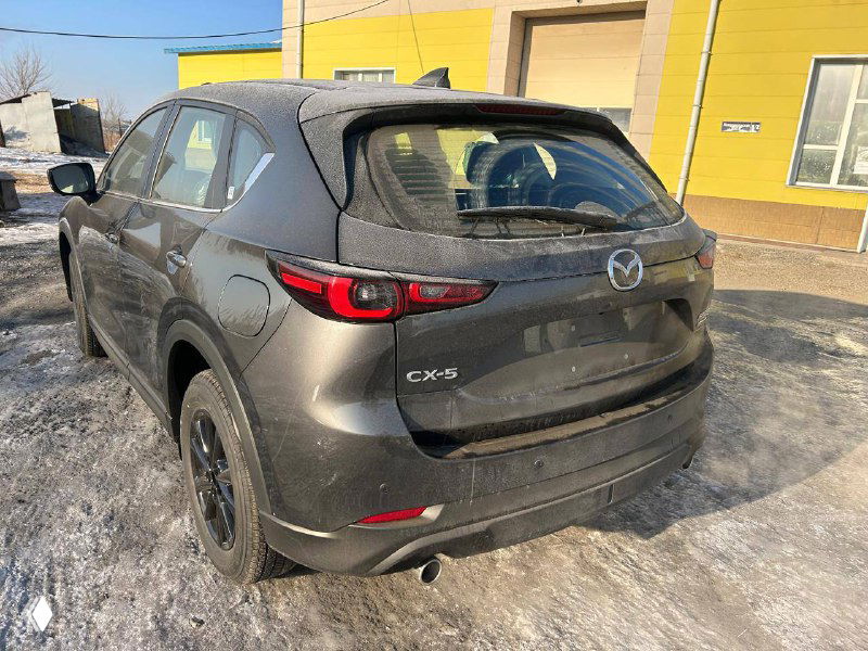 Задний вид Mazda CX-5 2025 года на складе: багажная дверь, фонари и эмблема CX-5, автомобиль припаркован на обледенелой площадке рядом с ангаром.