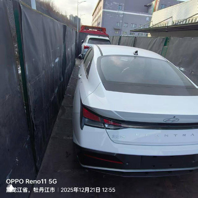 Вид сзади белой Hyundai Elantra в узком коридоре между ограждениями, видно заднюю часть и багажную крышку.