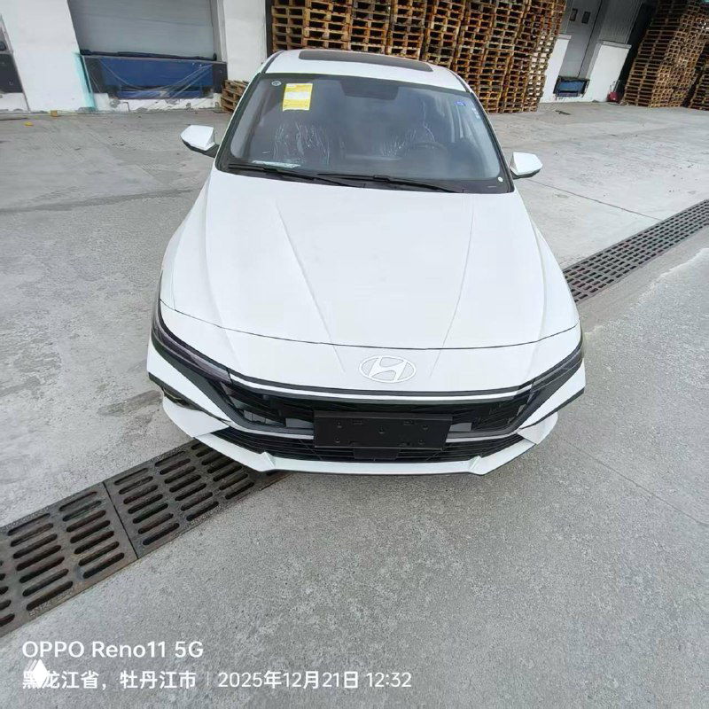 Фронтальный вид белой Hyundai Elantra на площадке, видно передний бампер, фары и наклейку на лобовом стекле.