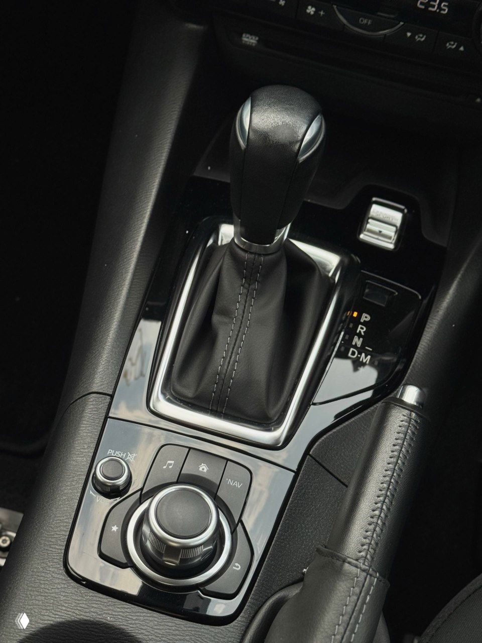 Блок рычага АКПП и органы управления мультимедиа Mazda Axela — крупный план селектора передач, ретро-панели и управляющего колеса центральной консоли.