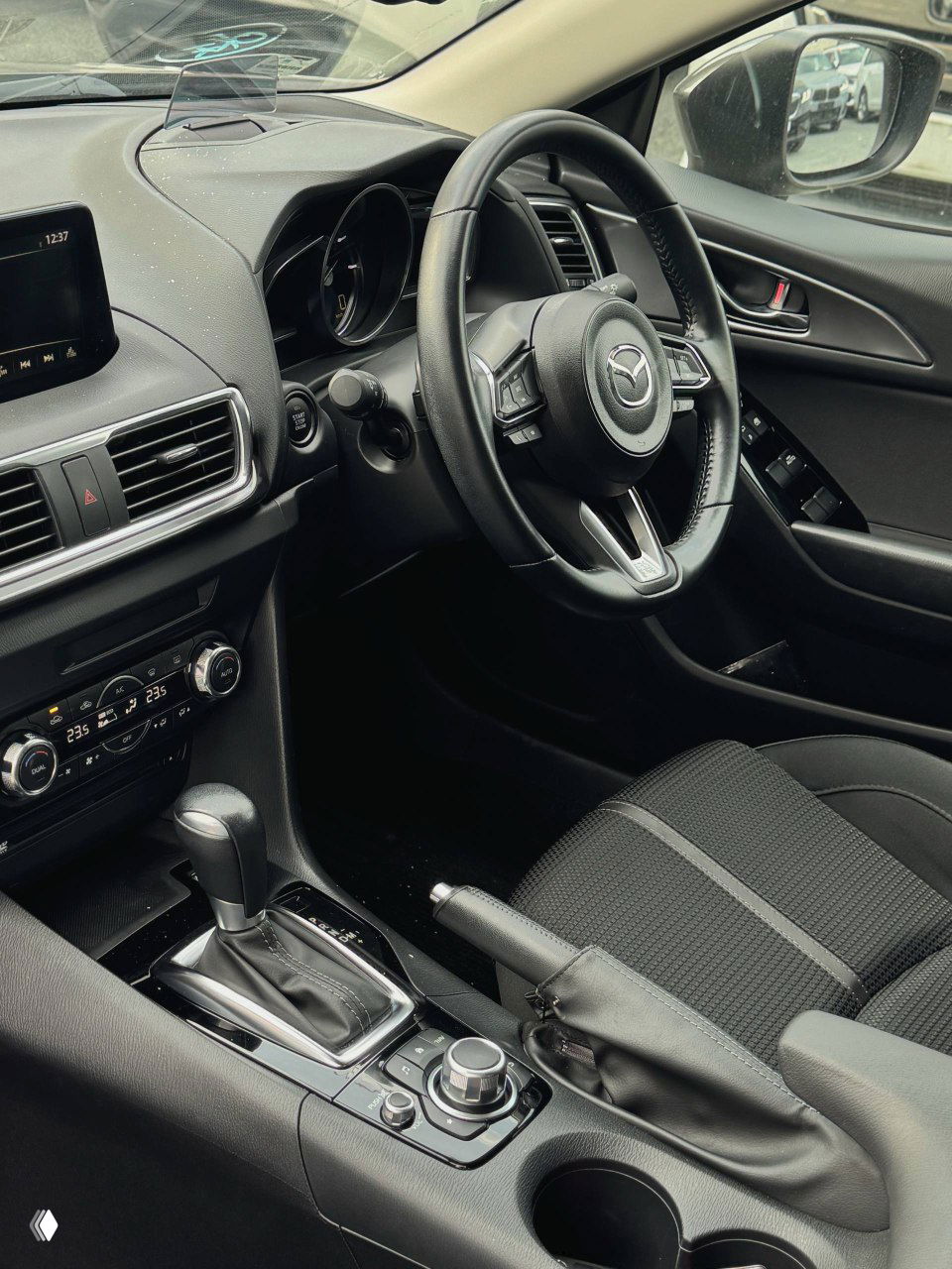 Интерьер Mazda Axela — вид на передние сиденья, рычаг ручного тормоза, подстаканники и центральную консоль, тканевая обивка и чистое состояние салона.