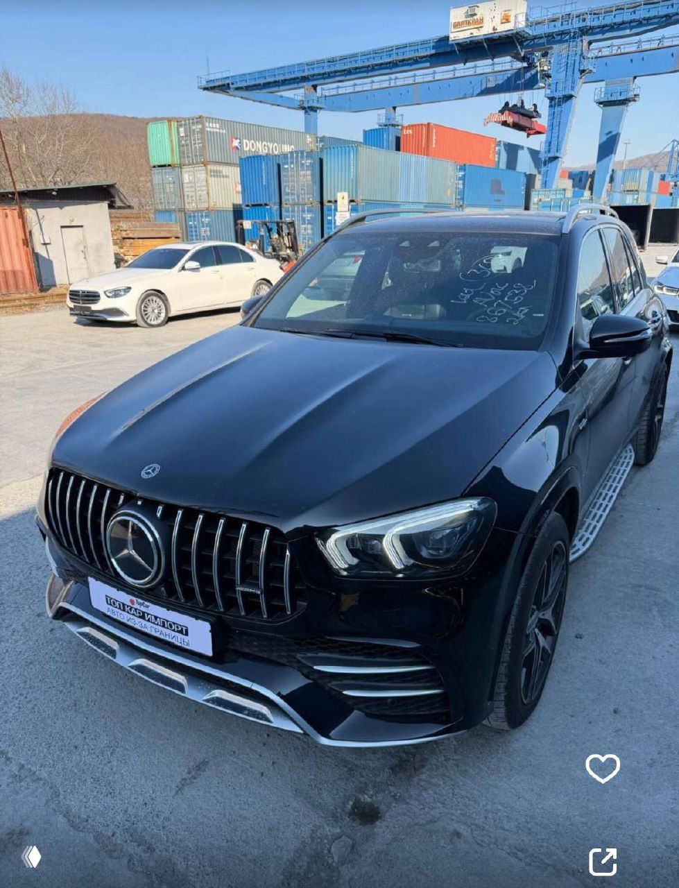 Чёрный Mercedes 53AMG на стоянке в порту с контейнерами и краном на заднем плане; вид спереди-углом, логотип и номер с брендом компании.