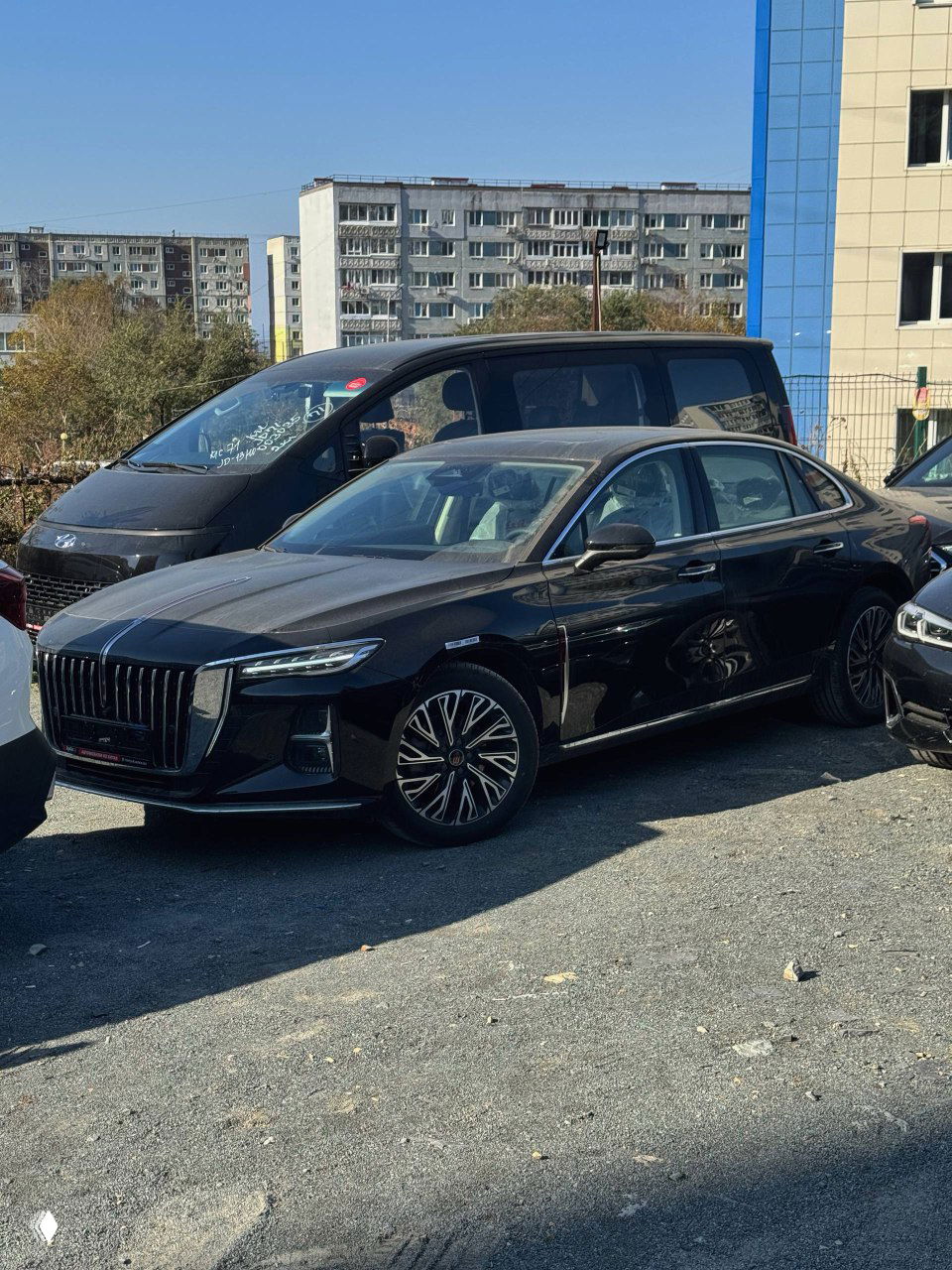 Общий вид черного Hongqi H5 на парковке рядом с другими автомобилями, вид сбоку под углом, отражения на кузове при дневном свете.