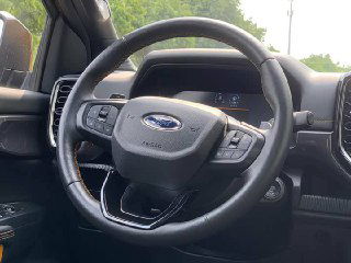 Крупный план рулевого колеса Ford Ranger с логотипом, видна приборная панель, кнопки управления и чёрная отделка рулевой колонки, фото салона.