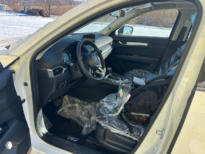 Интерьер Mazda CX-5 2025: передние кресла в защитной пленке, руль и центральная консоль; снимок сделан с открытой водительской двери.