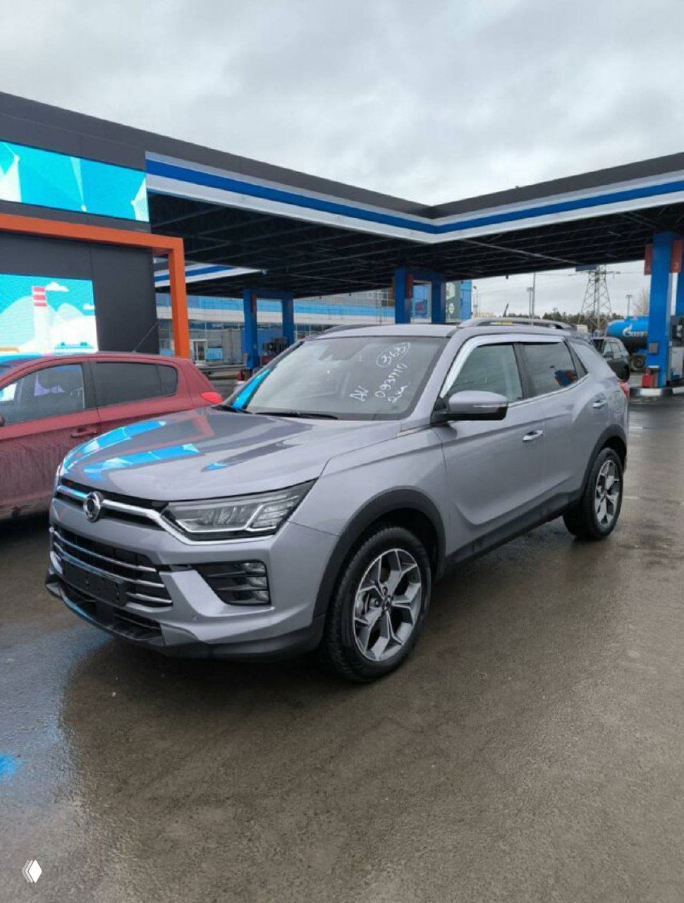 Крупный план серебристого SsangYong Korando у заправочной/портовой площадки, автомобиль на мокрой поверхности — изображение из отчёта о покупке.