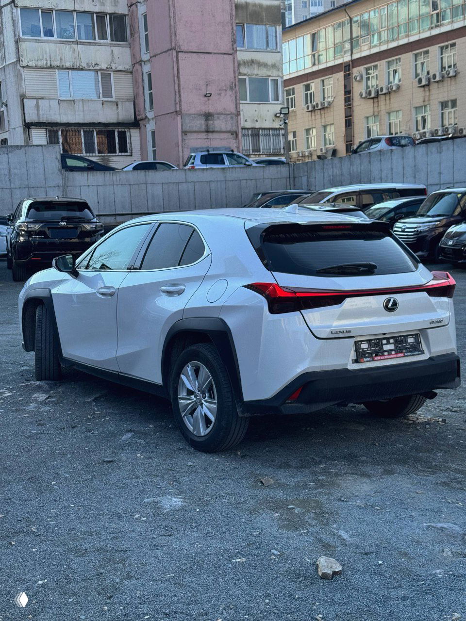 Белый Lexus UX200, вид сзади и в профиль на парковке; видно боковую часть, задние фонари и номерной подрамник без регистрации.