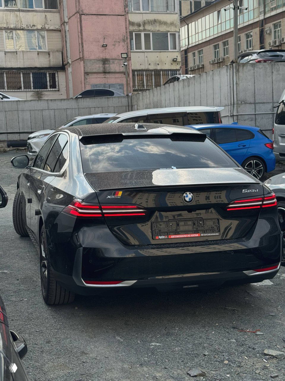 Задняя часть BMW 523d с заметными задними фонарями, закрытым номерным рамком и наклейками на багажной двери на стоянке.
