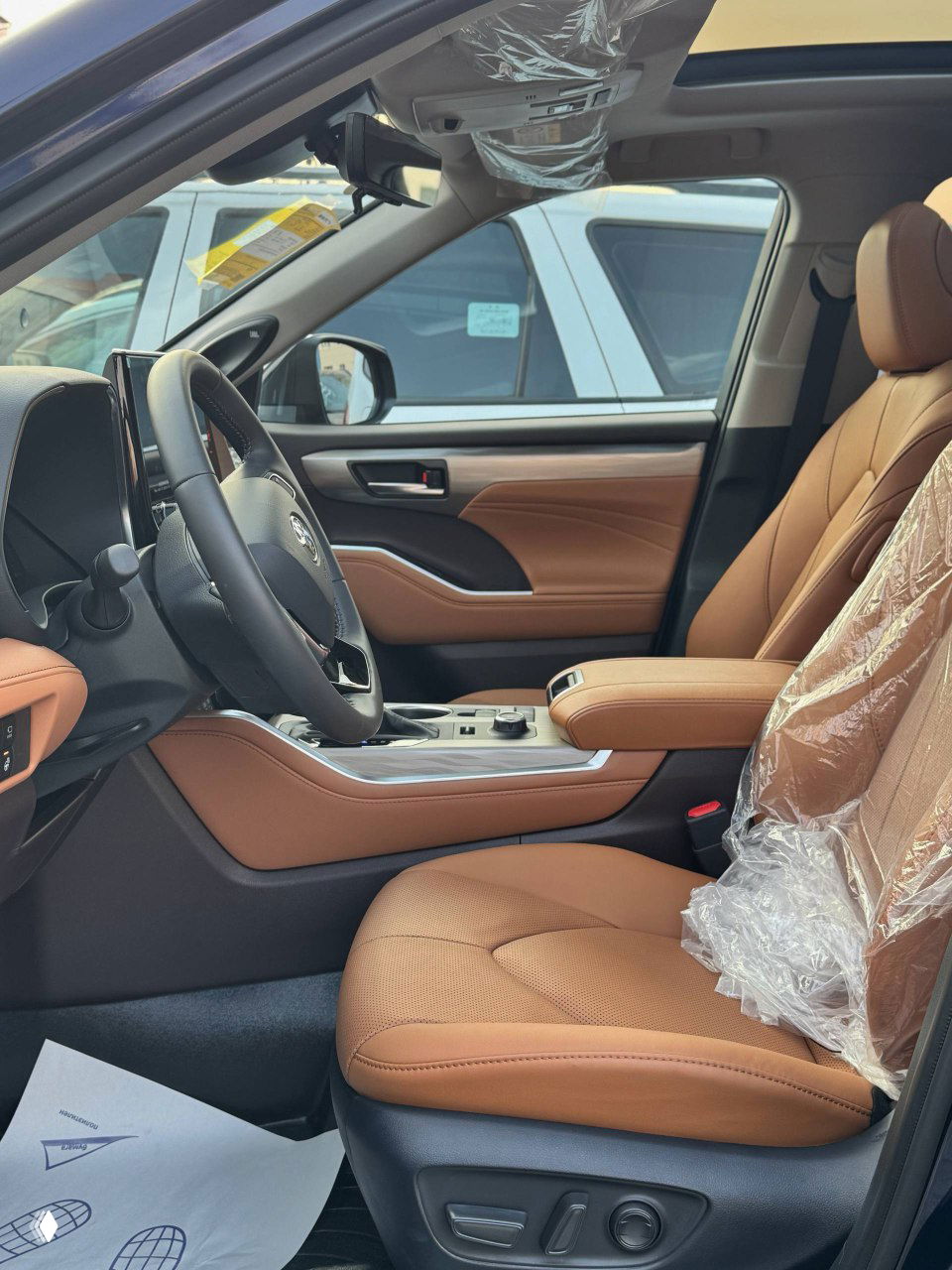 Водительское кресло и передняя панель Toyota Highlander 2025 в коричневой коже, защитная плёнка на кресле и наклейки на стекле свидетельствуют о новой поставке