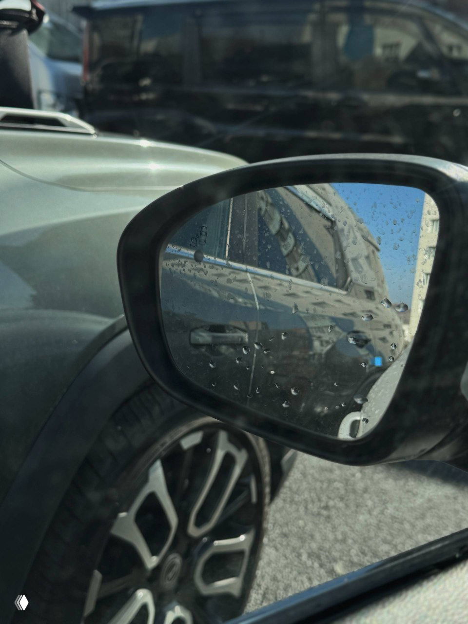Боковое зеркало и отражение соседнего автомобиля на фоне колесного диска Renault Samsung XM3; капли воды на стекле и детали зеркала.
