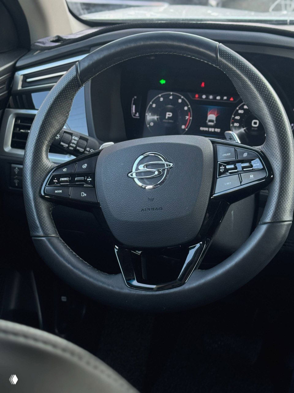 Рулевое колесо SsangYong с логотипом на коже, крупный план панели управления на руле и комбинированная приборная панель на заднем плане.