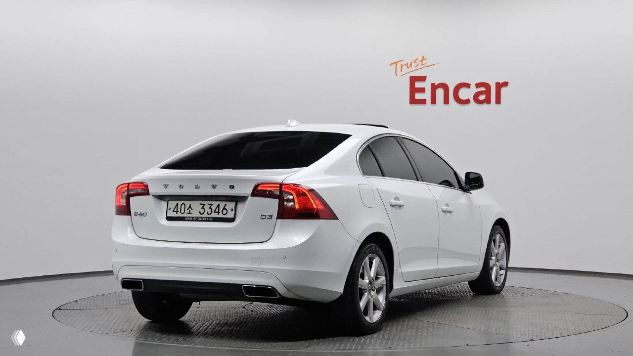 Вид сзади-лево белого Volvo S60 с корейским номером на подиуме Encar, видна задняя оптика и спортивный бампер автомобиля.
