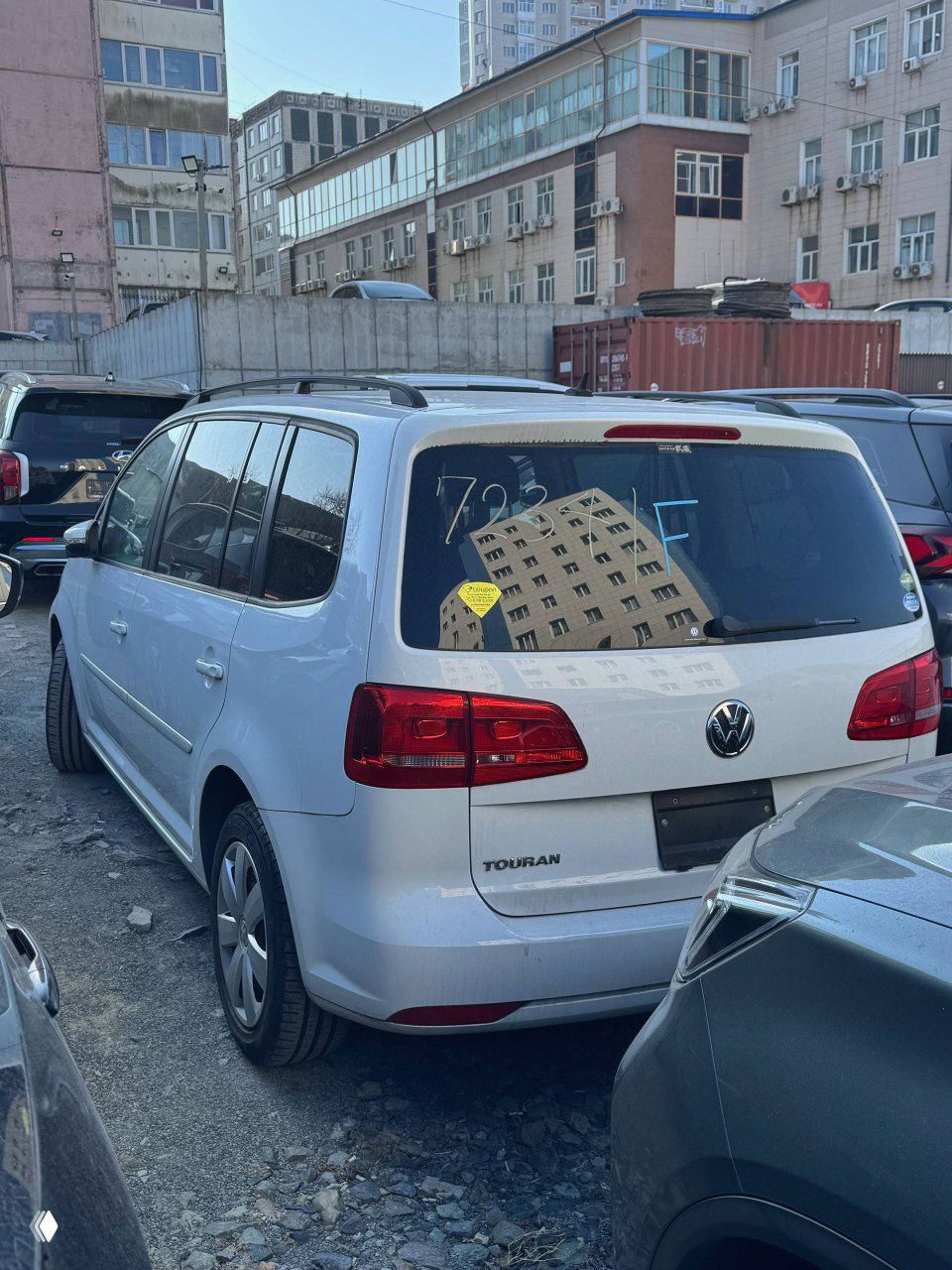 Задняя часть Volkswagen Touran 2014 на стоянке: эмблема VW, надпись TOURAN на багажнике, стекло с отражением зданий и парковочных мест.