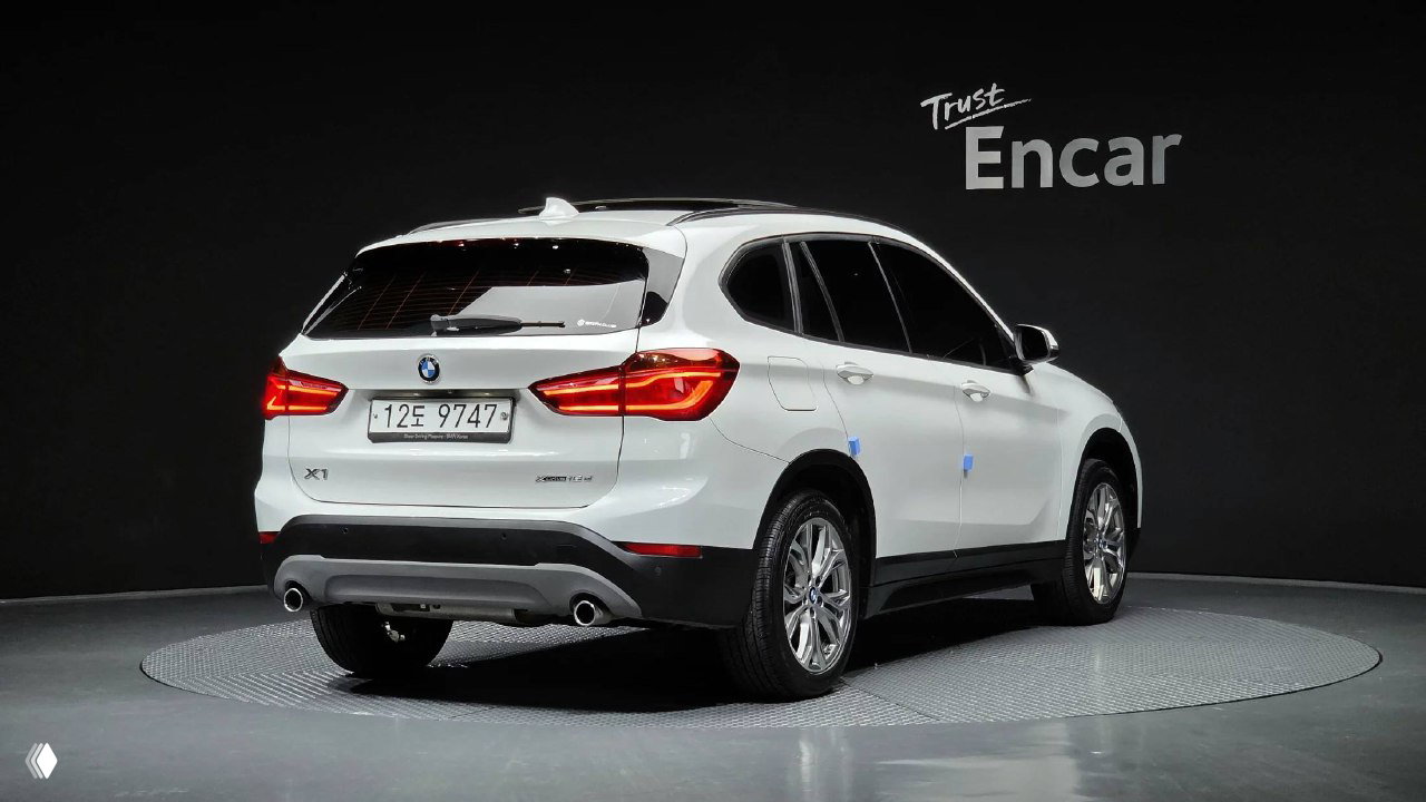 Вид сзади и сбоку белого BMW X1 xDrive 18d на подиуме шоурума, видно задние фонари, бампер и корейский номерной знак автомобиля.