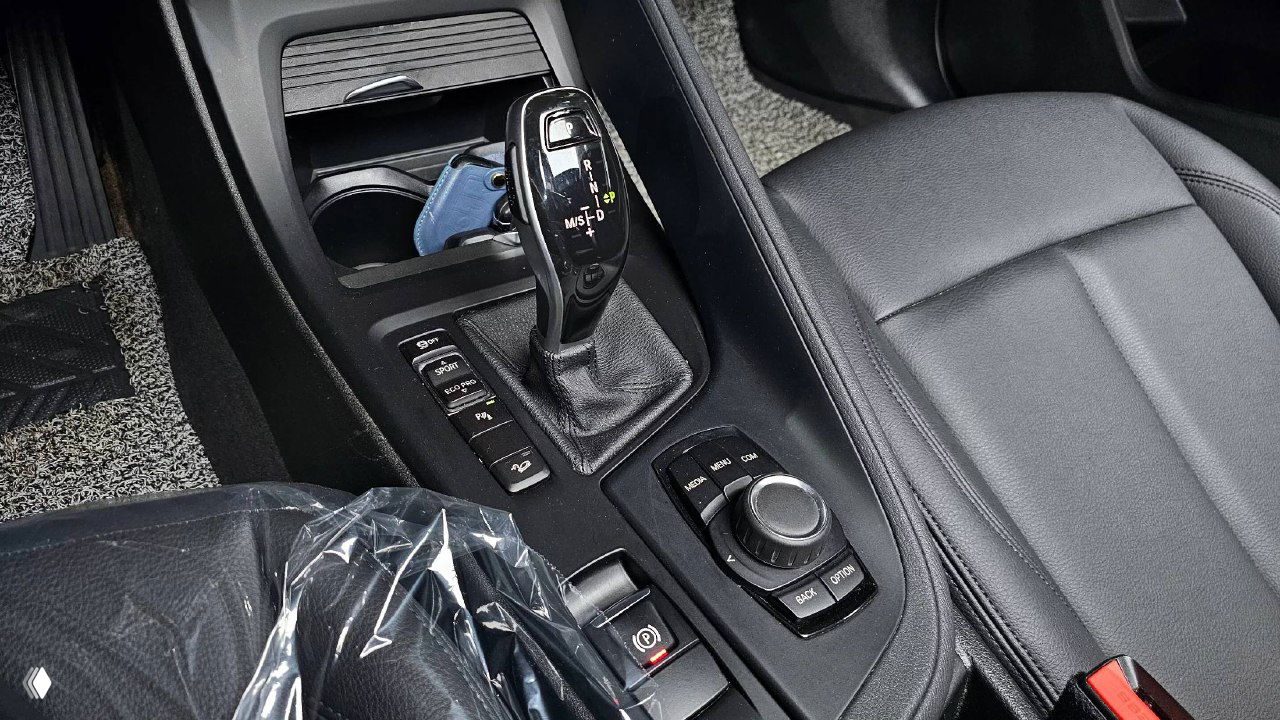 Селектор передач, кнопки режимов и контроллер iDrive на центральной консоли BMW X1, видна отделка и подстаканники рядом.