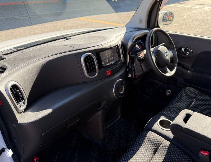Салон Nissan Cube: панель приборов, рулевое колесо и передние сиденья, вид в дневном освещении, состояние обивки и торпедо видно отчётливо.